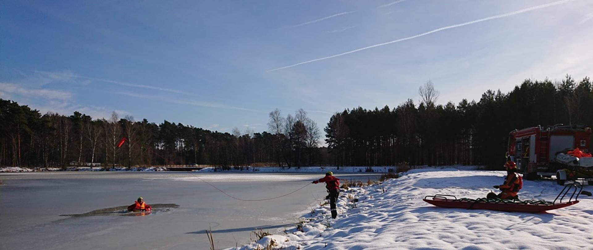 Na zdjęciu widzimy akwen wodny pokryty lodem. W wyniku zarwania się lodu w wodzie znajduje się poszkodowana osoba. Na brzegu zbiornika w bezpiecznej od niego odległości znajduje się samochód ratowniczo-gaśniczy oraz przyczepka do ratownictwa wodno-lodowego. Jeden ze strażaków stojący na brzegu wykonuje rzut rzutką ratowniczą w kierunku osoby znajdującej się w wodzie. Drugi za pomocą linki ratowniczej asekuruje strażaka podejmującego próbę udzielenia pomocy poszkodowanemu. Obok strażaka zajmującego się asekuracją znajdują się sanie lodowe. Strażacy ubrani są w ubrania wypornościowe, kamizelki ratunkowe..