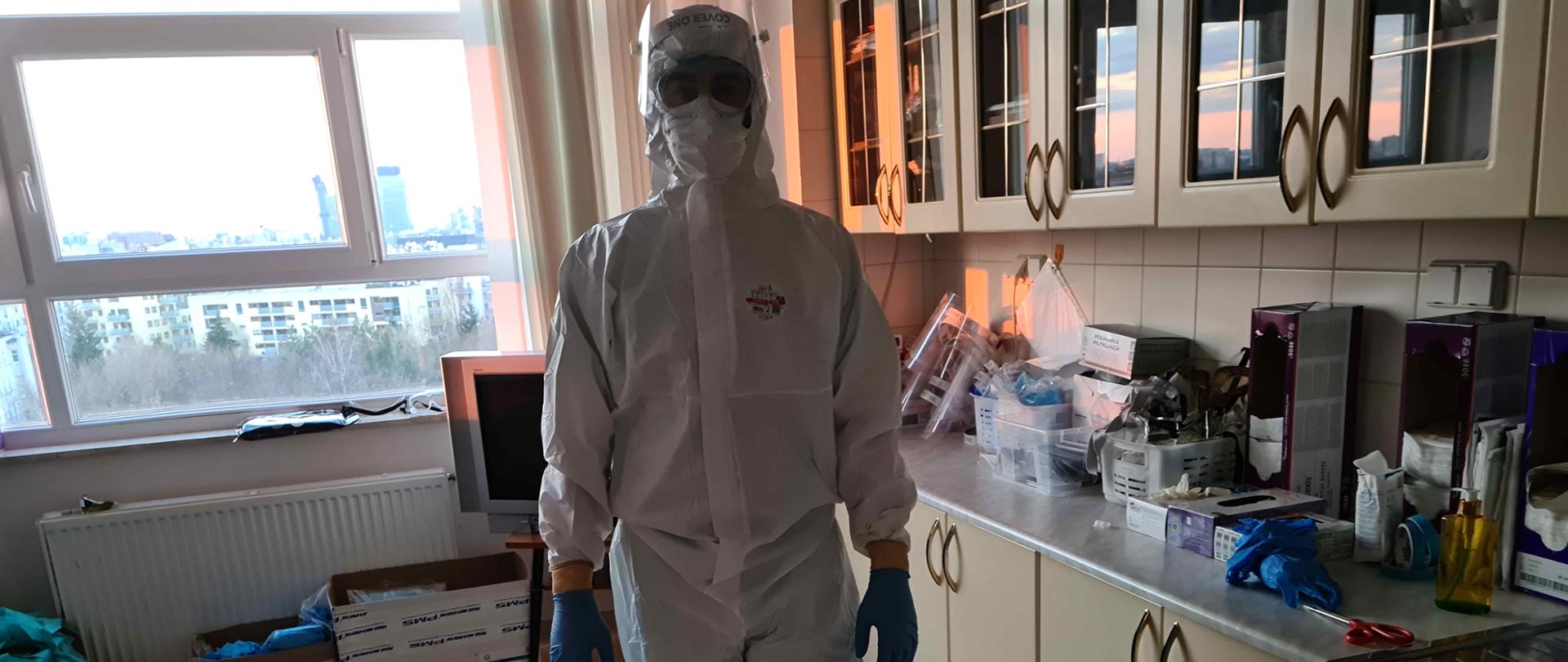 Zdjęcie zrobione w pomieszczeniu szpitala. Zdjęcie przedstawia stojącego strażaka ratownika medycznego, który jest ubrany w kombinezon ochronny, maskę, przyłbicę, gogle, rękawice lateksowe. Strażak jest gotowy do pomocy osoba chorym na COVID-19. W oddali widać poukładane na szafkach środki ochrony indywidualnej. 