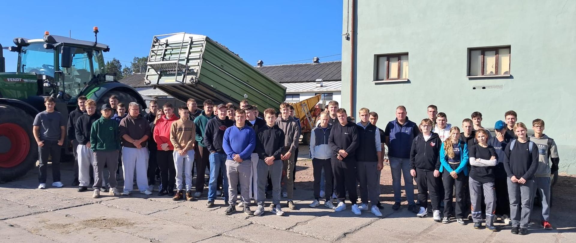 Grupa uczniów pozuje do zdjęcia na podwórzu gospodarstwa, obok zielonego ciągnika i podniesionej przyczepy z ziemniakami.