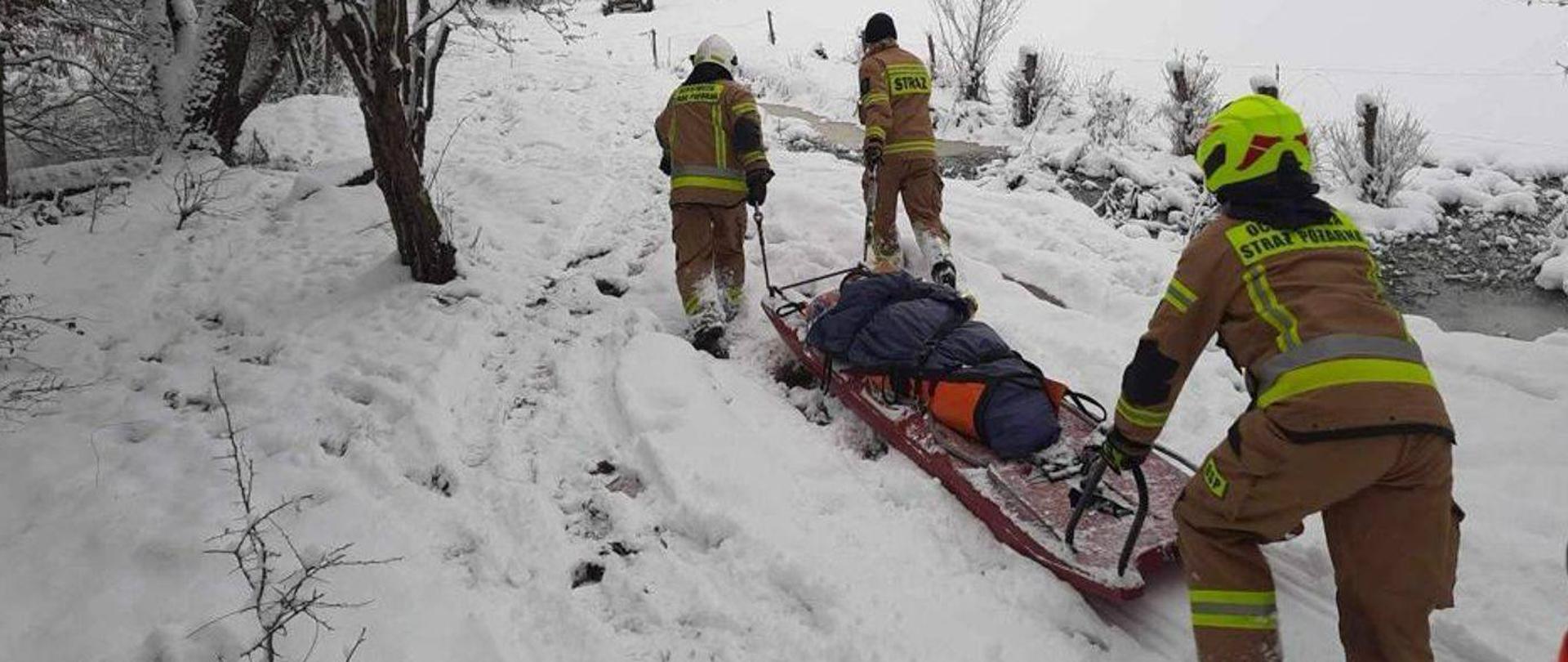 Zima, dużo śniegu. Strażacy transportują chorego do karetki na saniach.