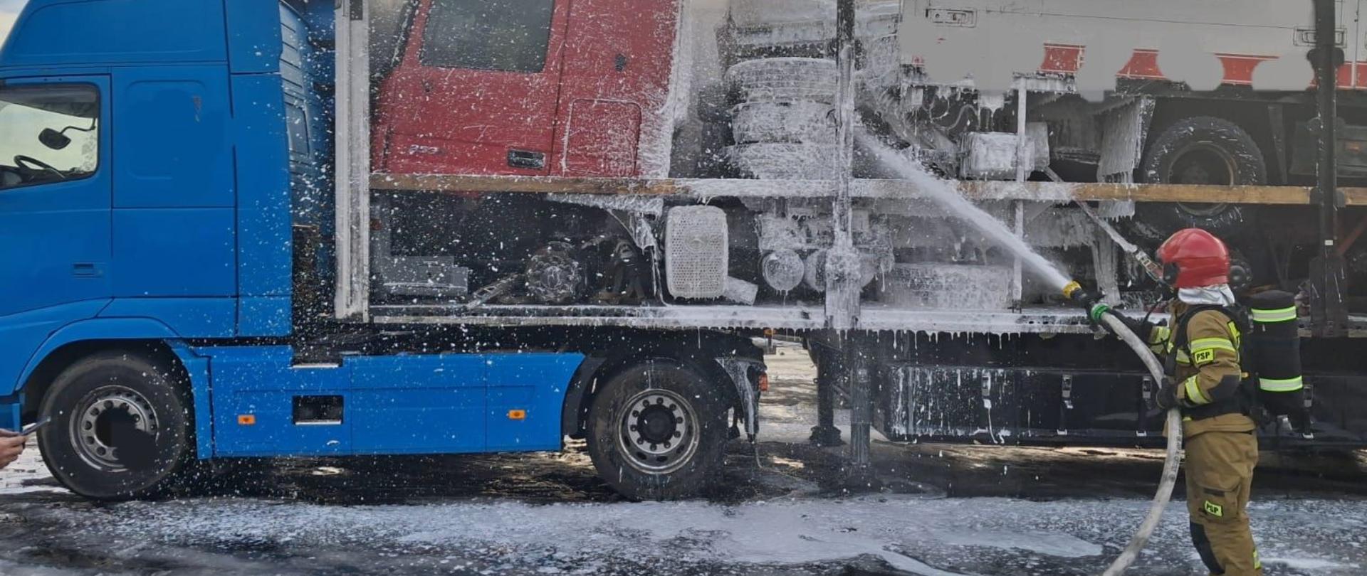 Na zdjęciu widać akcję gaśniczą. Po prawej stronie stoi strażak w pełnym umundurowaniu ochronnym i hełmie, który przy użyciu węża strażackiego rozprowadza pianę gaśniczą. Piana pokrywa konstrukcję naczepy oraz znajdujący się na niej samochód ciężarowy. Podłoże jest mokre i pokryte pianą oraz lodem lub śniegiem.