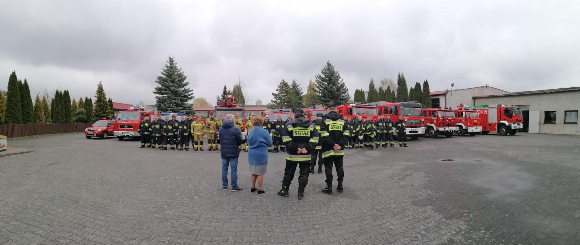 Podsumowanie ćwiczeń - ratownicy w dwuszeregu na tle samochodów strażackich. Tyłem stoją organizatorzy ćwiczeń