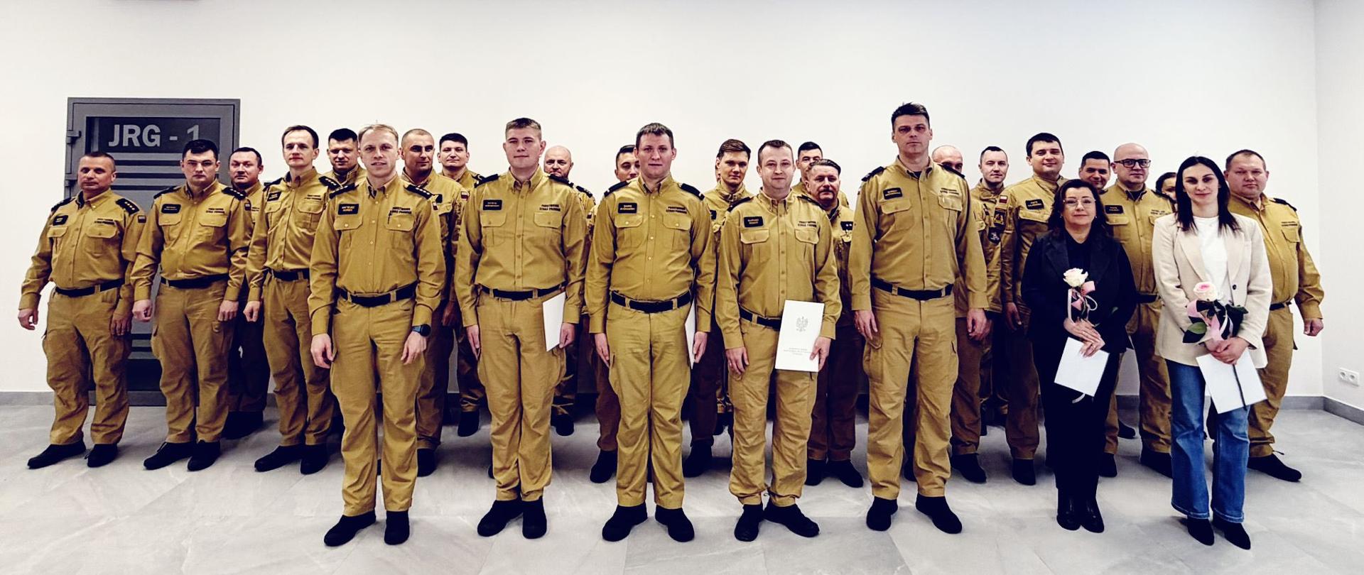 W jasnym, przestronnym pomieszczeniu stoi duża grupa osób ustawiona w kilku rzędach do wspólnej fotografii. Większość z nich to strażacy w jednakowych, beżowych mundurach i czarnych butach. Stoją prosto, blisko siebie, tworząc zwartą formację.
Z przodu, na środku, znajduje się kilka osób w cywilnych ubraniach — eleganckich koszulach i marynarkach. Jedna z kobiet trzyma bukiet kwiatów, a inna osoba trzyma dokumenty lub teczkę. Wygląda to na moment uroczysty.