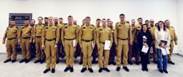 W jasnym, przestronnym pomieszczeniu stoi duża grupa osób ustawiona w kilku rzędach do wspólnej fotografii. Większość z nich to strażacy w jednakowych, beżowych mundurach i czarnych butach. Stoją prosto, blisko siebie, tworząc zwartą formację.
Z przodu, na środku, znajduje się kilka osób w cywilnych ubraniach — eleganckich koszulach i marynarkach. Jedna z kobiet trzyma bukiet kwiatów, a inna osoba trzyma dokumenty lub teczkę. Wygląda to na moment uroczysty