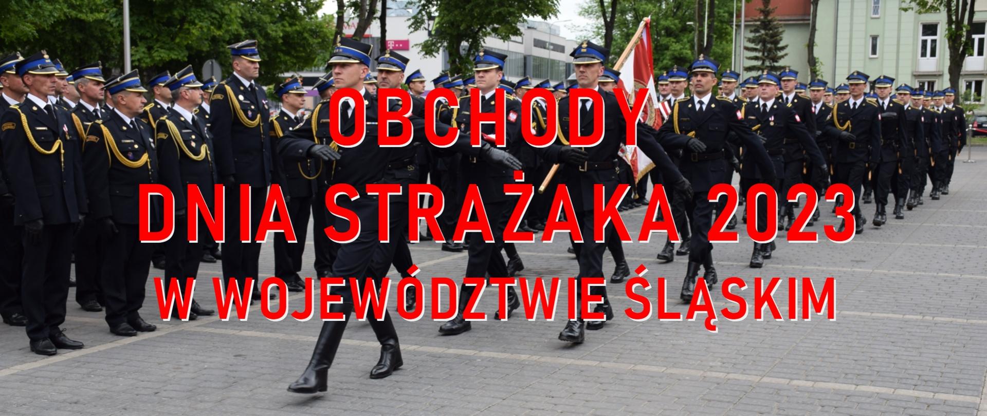 na zdjęciu widoczni strażacy w mundurach wyjściowych z komapanii reprezentacyjnej komendy wojewódzkiej Państwowej Straży Pożarnej w Katowicach. Strażacy maszeruja w szyku. Jako pierwszy dowódca kompanii za nim strażacy z pocztu sztandarowego. Na zdjęciu widnieje napis w kolorze czerwonym Obchody Dnia Strażaka 2023 w województwie śląskim