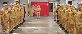 Strażacy stoją w garażu w dwóch szeregach zwróconych naprzeciwko siebie. Na głównym planie stoi komendant miejski Państwowej Straży Pożarnej w Świętochłowicach, który przemawia do zebranych strażaków.