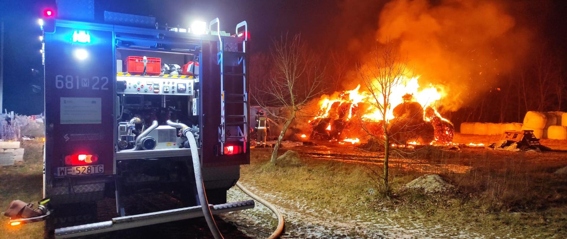 Na zdjęciu na pierwszym planie po lewej stronie widzimy tył wozu strażackiego pochodzący z KP PSP Wołomin, filia Tłuszcz. Samochód ma włączone sygnały świetlne barwy niebieskiej. Widzimy odsłoniętą tylną skrytkę w pojeździe, w której znajduje się autopompa. Po prawej stronie na zdjęciu widzimy składowisko bel słomy, które są objęte pożarem. Miedzy samochodem, a belami widać strażaków, ubranych w ubranie specjalne typu Nomex, którzy podają jedne prąd wody na pożar. Po prawej stronie pożaru widać kolejne składowisko, są to ustawione bele kiszonki.