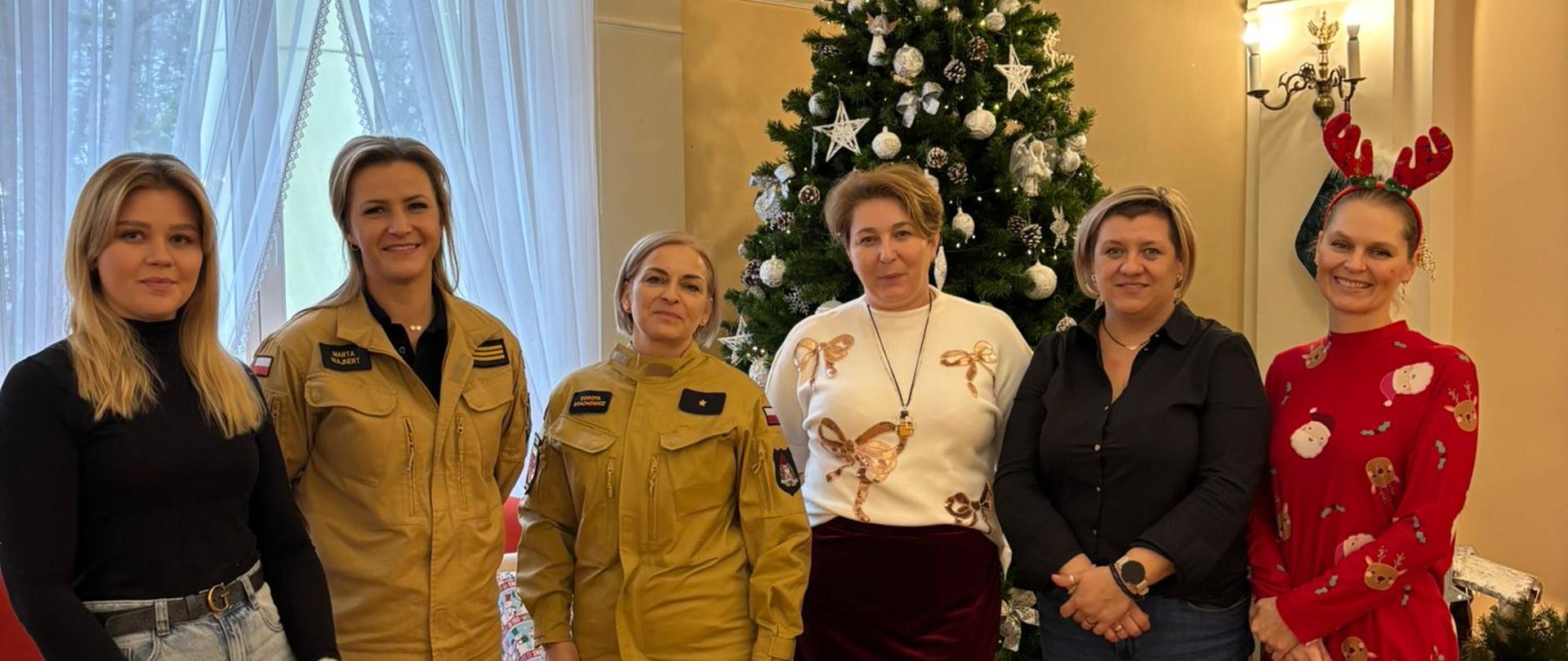 Grupa sześciu kobiet stoi w eleganckiej sali przed udekorowaną choinką bożonarodzeniową. Dwie z nich ubrane są w umundurowanie Państwowej Straży Pożarnej. Pozostałe kobiety ubrane są w stroje codzienne. Jedna z pań ma ubranie w świątecznym motywie. W tle widoczne prezenty świąteczne ułożone pod choinką oraz jasne, wysokie okna, tworzące ciepłą, świąteczną atmosferę. 
