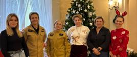 Grupa sześciu kobiet stoi w eleganckiej sali przed udekorowaną choinką bożonarodzeniową. Dwie z nich ubrane są w umundurowanie Państwowej Straży Pożarnej. Pozostałe kobiety ubrane są w stroje codzienne. Jedna z pań ma ubranie w świątecznym motywie. W tle widoczne prezenty świąteczne ułożone pod choinką oraz jasne, wysokie okna, tworzące ciepłą, świąteczną atmosferę. 