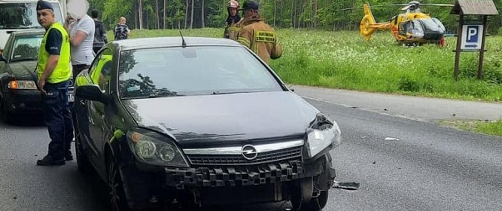 Zdjęcie przedstawia widoczny od przodu uszkodzony pojazd marki Opel z uszkodzoną lampą przednia od strony kierowcy oraz oderwanym zderzakiem przednim. Po prawej stronie stojący na łące żółty śmigłowiec LPR. Za wrakiem pojazdu strażacy i policjanci w umundurowaniu