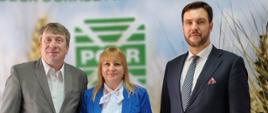 Trzy osoby pozują do zdjęcia na tle tego samego baneru z napisem „Podkarpacki Ośrodek Doradztwa Rolniczego w Boguchwale” i motywem zbóż. Po lewej stoi mężczyzna w jasnoszarym garniturze i białej koszuli, uśmiechnięty. W środku kobieta w niebieskim garniturze i białej bluzce z kokardą, trzymająca czarną torebkę, również lekko się uśmiecha. Po prawej mężczyzna w ciemnym garniturze z krawatem i poszetką, stojący prosto z neutralnym wyrazem twarzy. Wszyscy wyglądają elegancko i formalnie.