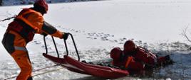 Na zdjęciu widoczni trzej strażacy na skutym lodzie akwenie wodnym. Wszyscy w suchych skafandrach koloru pomarańczowego w czerwonych kaskach ochronnych. Dawaj ratownicy z użyciem sań lodowych ewakuują z wody strażaka.