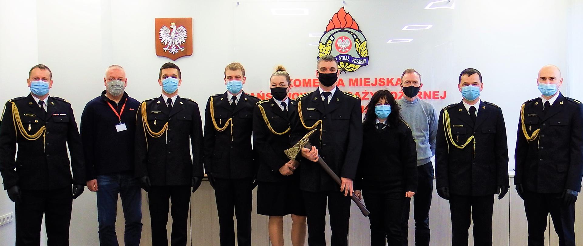 10 osób, w tym 8 w mundurach na tle godła państwowego i godła PSP. Po środku Sławomir Kulesza trzymający pamiątkowy topór strażacki.