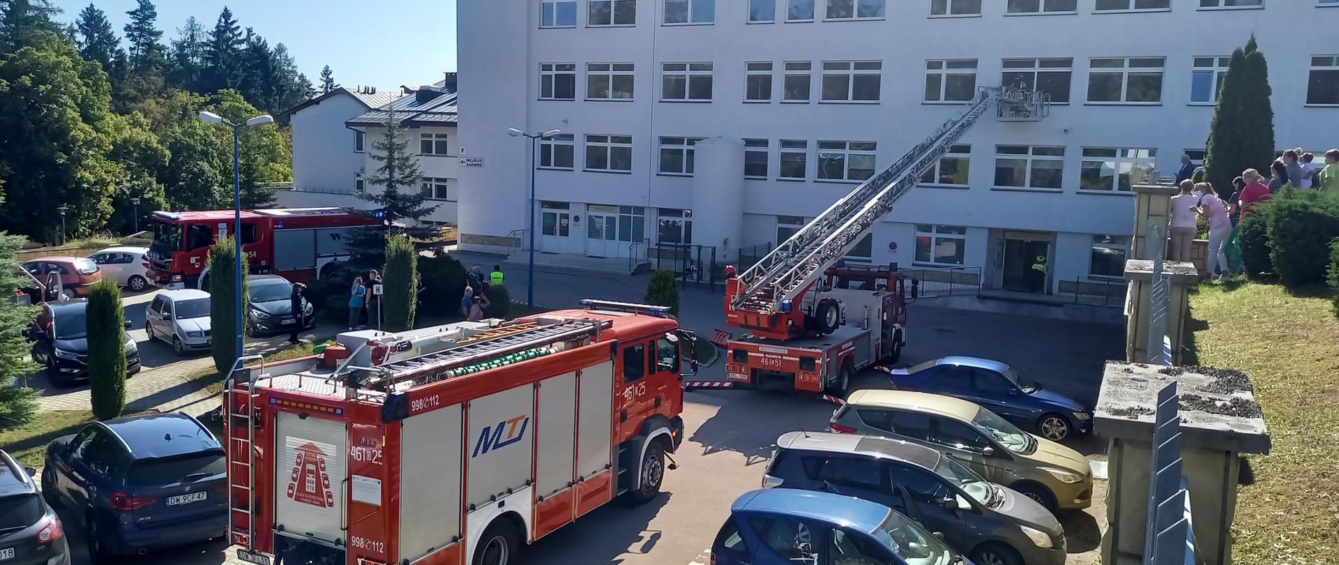 Widok na budynek szpitala. Przed nim samochód podnośnik z wysuniętym koszem, w którym znajduję się strażak prowadzący ewakuację przez okno.