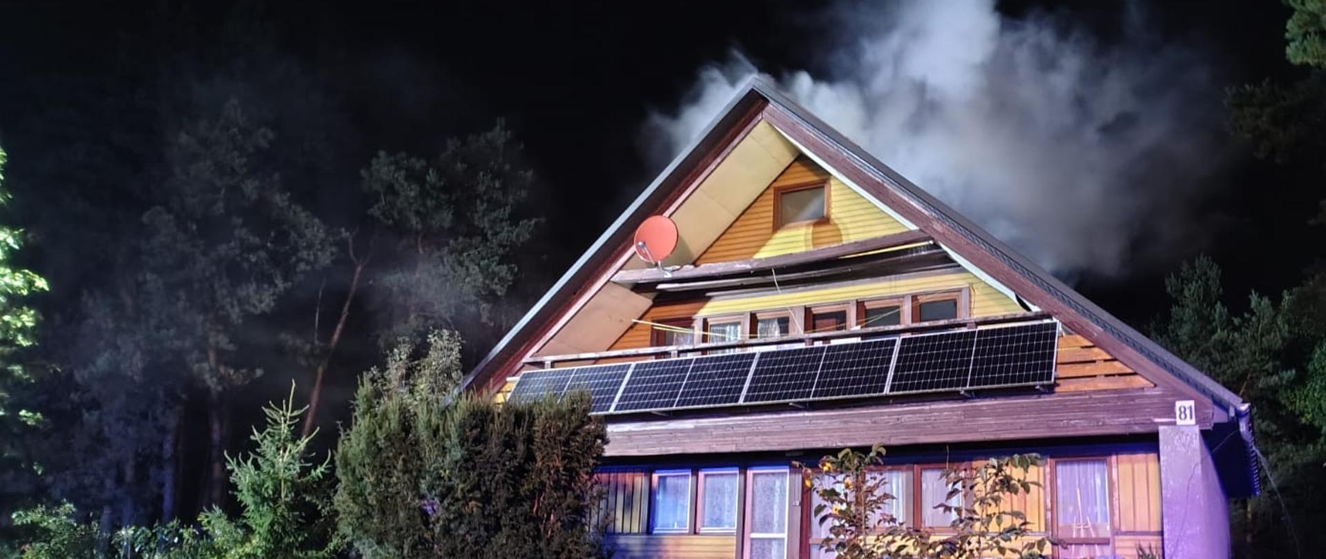 Zdjęcie przedstawia budynek jednorodzinny, gdzie doszło do pożaru. Nad dachem unosi się dym pożarowy i para wodna.