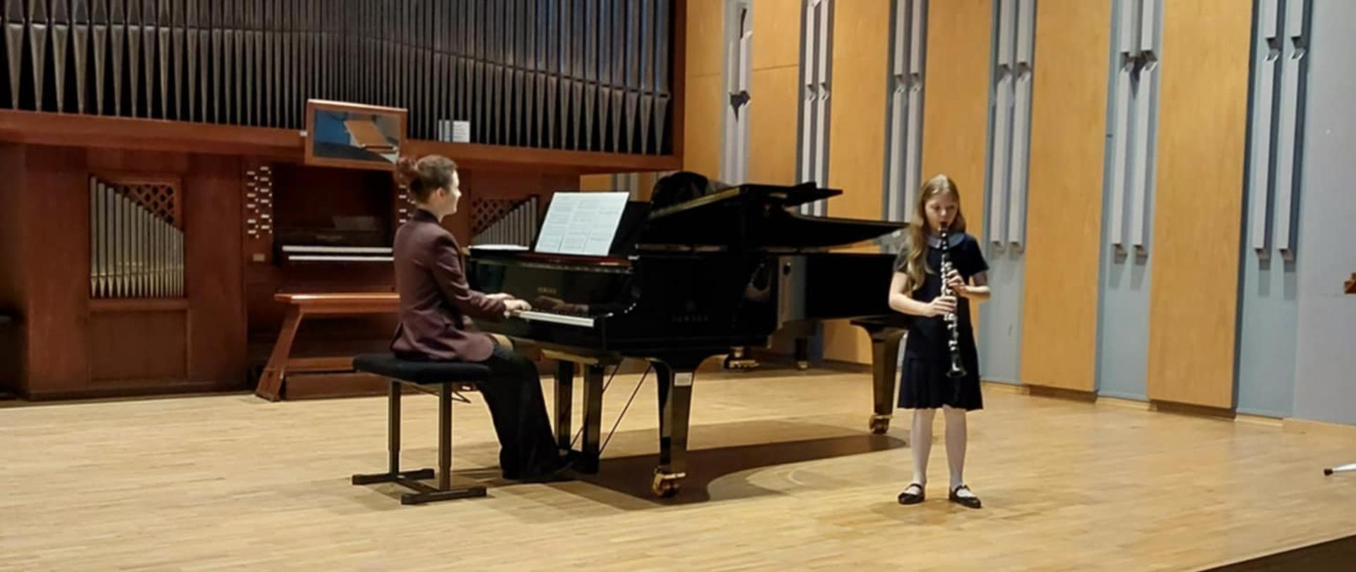 Na scenie sali koncertowej uczennica gra na klarnecie, obok niej na fortepianie gra akompaniatorka. Na pierwszym planie puste fotele. Za nimi organy oraz ściana sali koncertowej z kinkietami, na suficie zapalone lampy.