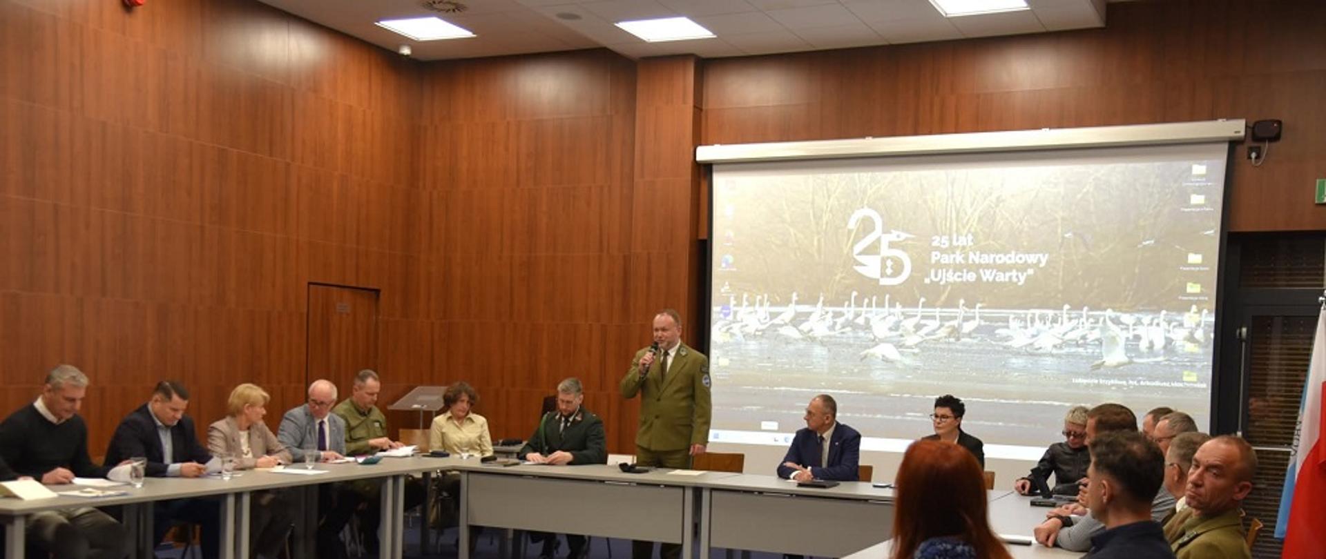 Przy stołach ustawionych w kształt podkowy siedzi kilkanaście osób. Mężczyzna w zielonym mundurze stoi i mówi do mikrofonu.