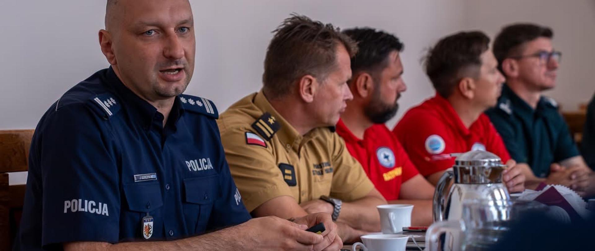 Pięciu mężczyzn w różnych mundurach służb ratunkowych i mundurowych siedzi przy stole konferencyjnym. Na pierwszym planie po lewej stronie przemawia policjant w ciemnoniebieskiej koszuli z napisem POLICJA. Obok niego siedzą kolejno strażak w piaskowym mundurze, dwaj ratownicy w czerwonych koszulkach oraz mężczyzna w ciemnozielonym mundurze. Na stole stoją filiżanki i dzbanki.