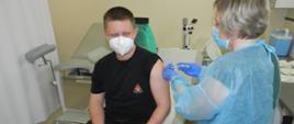 Na zdjęciu widoczny strażak w trakcie szczepienia przeciwko COVID-19 w ramach akcji #SzczepimySię Narodowego Programu Szczepień.