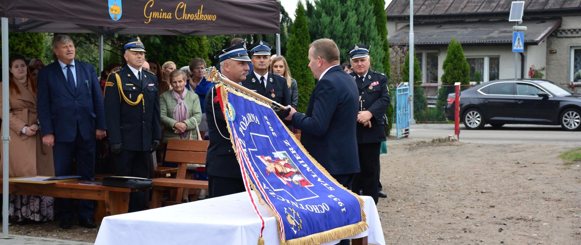 Przewodniczący komitetu społecznego fundacji sztandaru podejmuje ze stołu sztandar i przekazuje go strażakowi. W tle obserwatorzy - goście. W dali widać samochód osobowy i fragment miejscowej zabudowy.