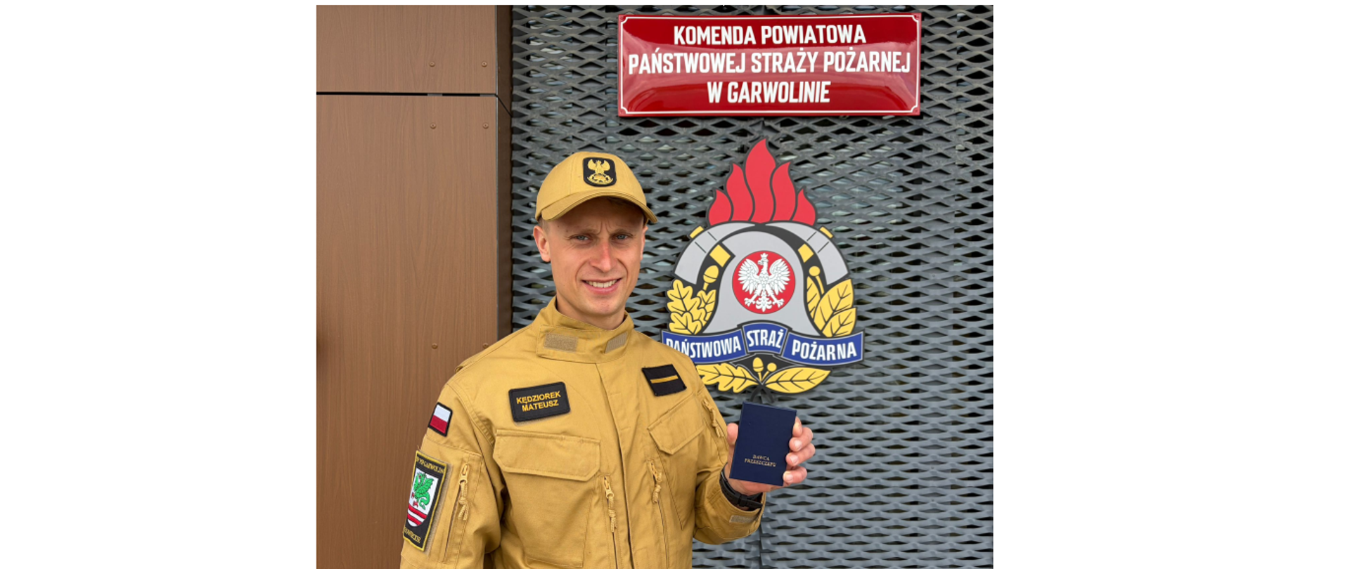 Zdjęcie przedstawia uśmiechniętego strażaka w piaskowym mundurze, który stoi przed budynkiem Komendy Powiatowej Państwowej Straży Pożarnej w Garwolinie. Mężczyzna prezentuje trzymaną w dłoni małą, niebieską legitymację z wyraźnym napisem „Dawca przeszczepu”. Na jego umundurowaniu widoczny jest imiennik „Kędziorek Mateusz”, a w tle umieszczono duże godło oraz czerwoną tablicę informacyjną formacji.