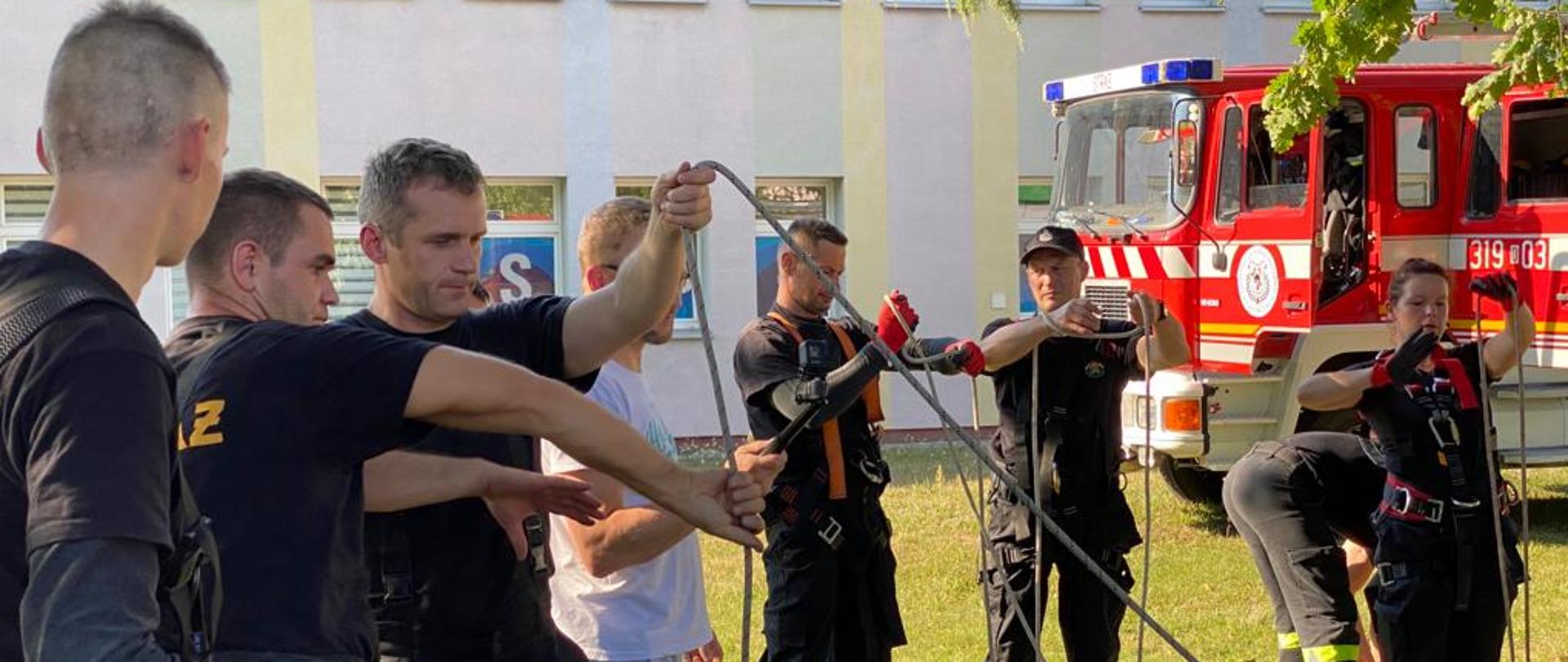 Strażacy OSP w ubraniu koszarowym uczą się wiązania węzłów na linach. Strażacy w trakcie szkolenia z ratownictwa wysokościowego w krajowym systemie ratowniczo-gaśniczym. W tle widoczny samochód pożarniczy koloru czerwonego.
