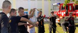 Strażacy OSP w ubraniu koszarowym uczą się wiązania węzłów na linach. Strażacy w trakcie szkolenia z ratownictwa wysokościowego w krajowym systemie ratowniczo-gaśniczym. W tle widoczny samochód pożarniczy koloru czerwonego.