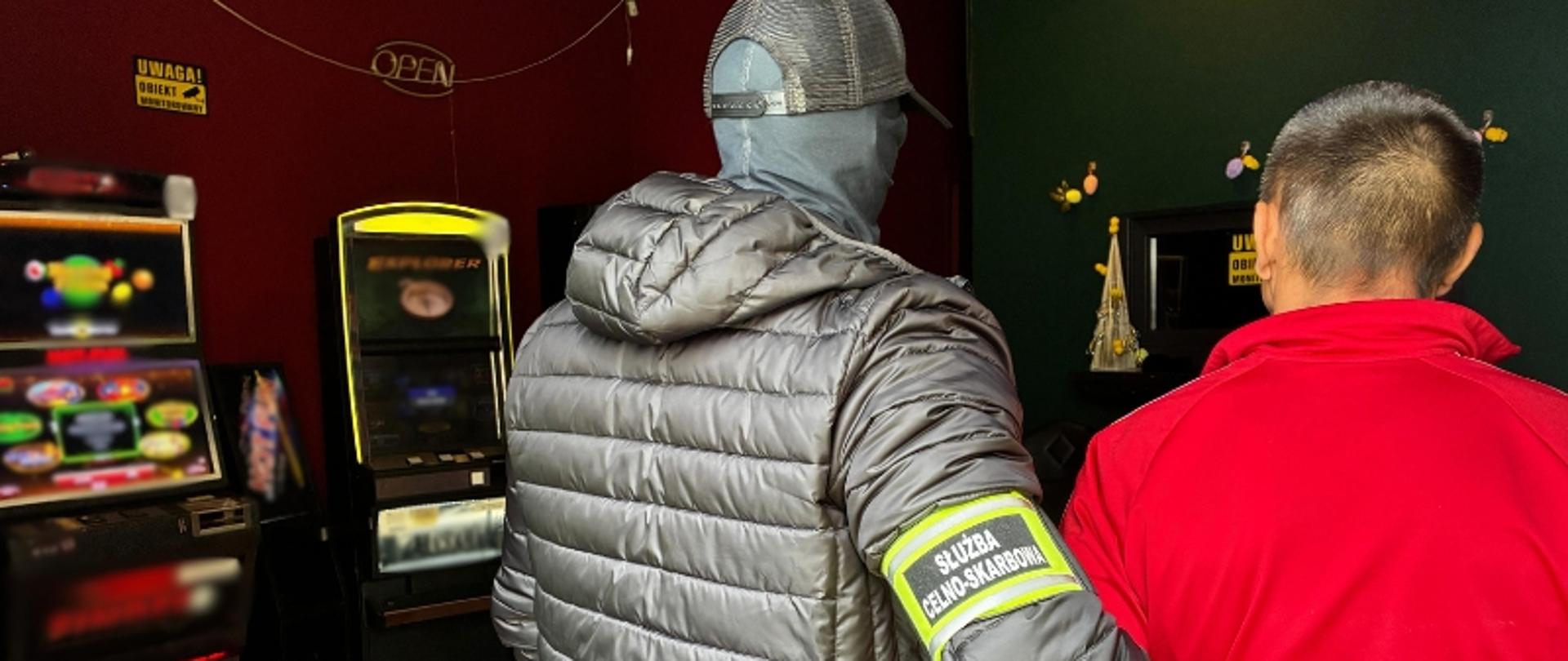 Funkcjonariusz Służby Celno-Skarbowej zatrzymuje podejrzanego o organizowanie nielegalnych gier hazardowych. Obok nielegalne automaty. 