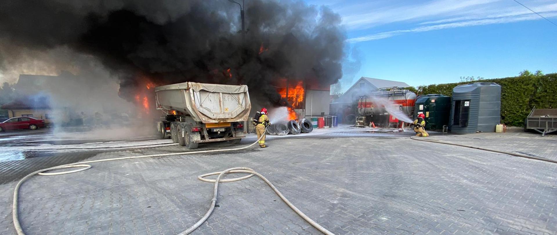 Zdjęcie przedstawia warsztat samochodowy, który w całości objęty jest płomieniami. Z budynku wydobywają się kłęby czarnego dymu. Przed garażem widać tył naczepy samochodu ciężarowego. Na placu utwardzonym kostką brukową rozciągnięte są węże strażackie. Dwóch strażaków trzymając w ręku prądownice wodne gasi pożar. 