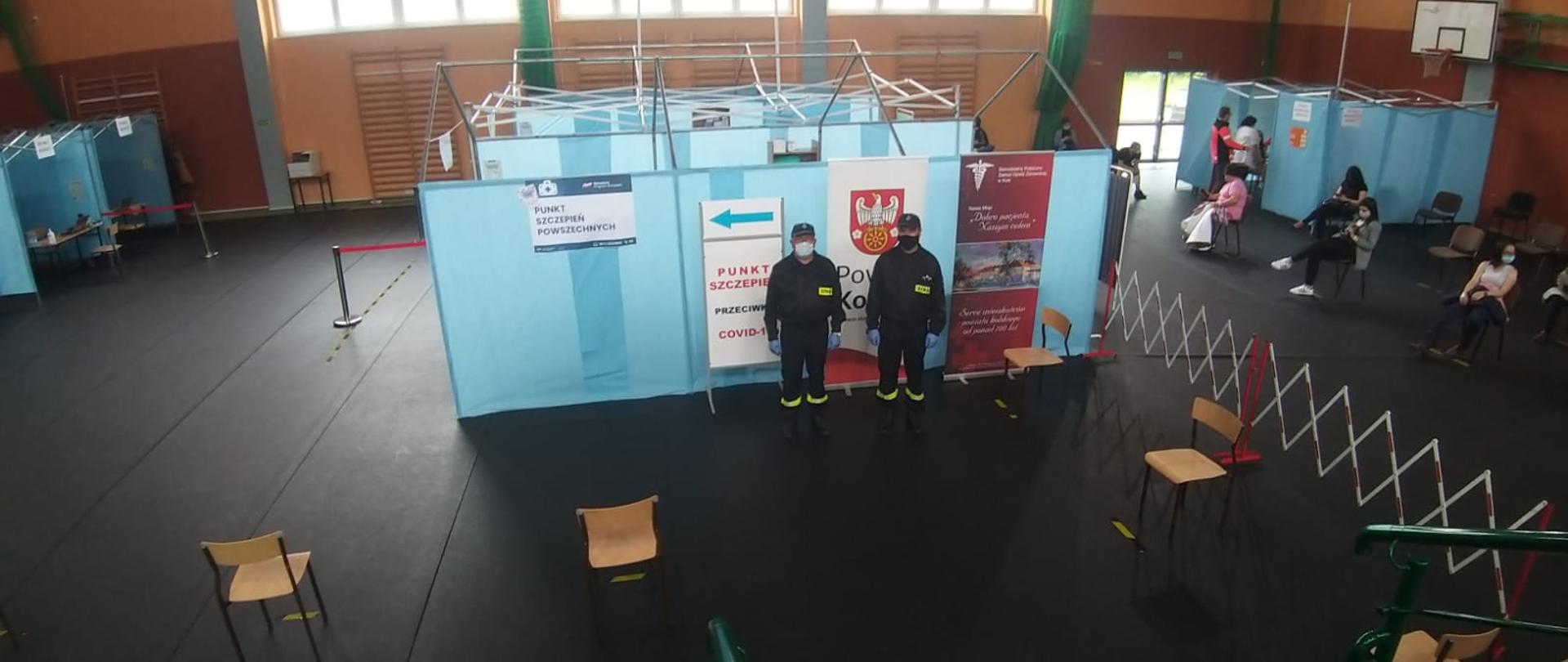 Zdjęcie przedstawia dwóch umundurowanych strażaków pomagających przy szczepieniach. Strażacy stoją na tle parawanów znajdujących się na terenie hali.