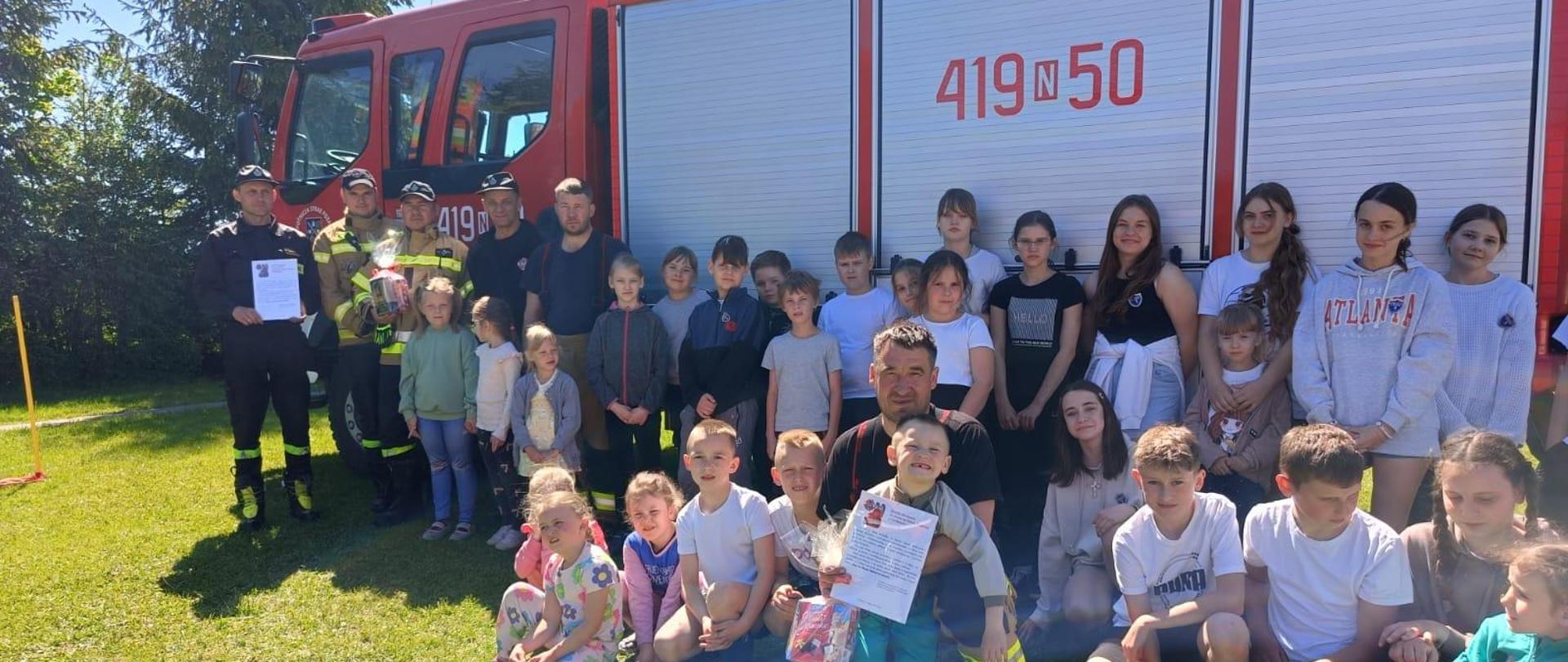Na zdjęciu duża grupa dzieci z przedszkola. Stoją ze strażakami na tle dużego samochodu strażackiego. Słonecznie.
