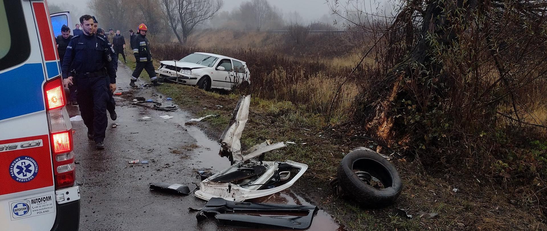 Na zdjęciu widać na pierwszym planie wrak oraz rozbite części samochodu osobowego VW Golf koloru białego. Z prawej strony drzewo przydrożne, w które wjechał samochód osobowy. W tle karetka pogotowia oraz policja i strażacy. Zdjęcie wykonane w porze zapadającego mroku.