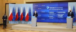Premier Matusz Morawiecki, wicepremier, minister rozwoju, pracy i technologii Jarosław Gowin stojący przy mównicach, w lewym rogu stoi rzecznik rządu Piotr Müller. Z tyłu za plecami premierów ekran z informacją o pomocy z tarczy branżowej dla rozszerzonej listy kodów