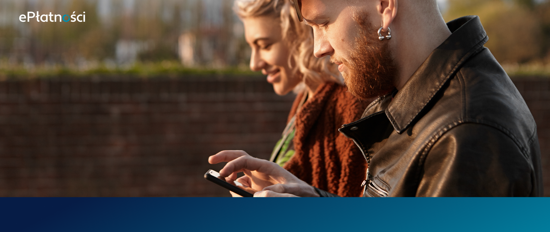 Na pierwszym planie brodaty mężczyzna z telefonem, obok niego uśmiechnięta kobieta. Ujęcie z boku.