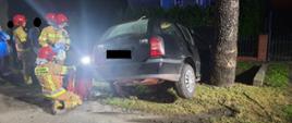 Widać ciemny uszkodzony samochód z prawej strony od tyłu, który jest blisko drzewa, jest noc, w pobliżu jest trzech strażaków