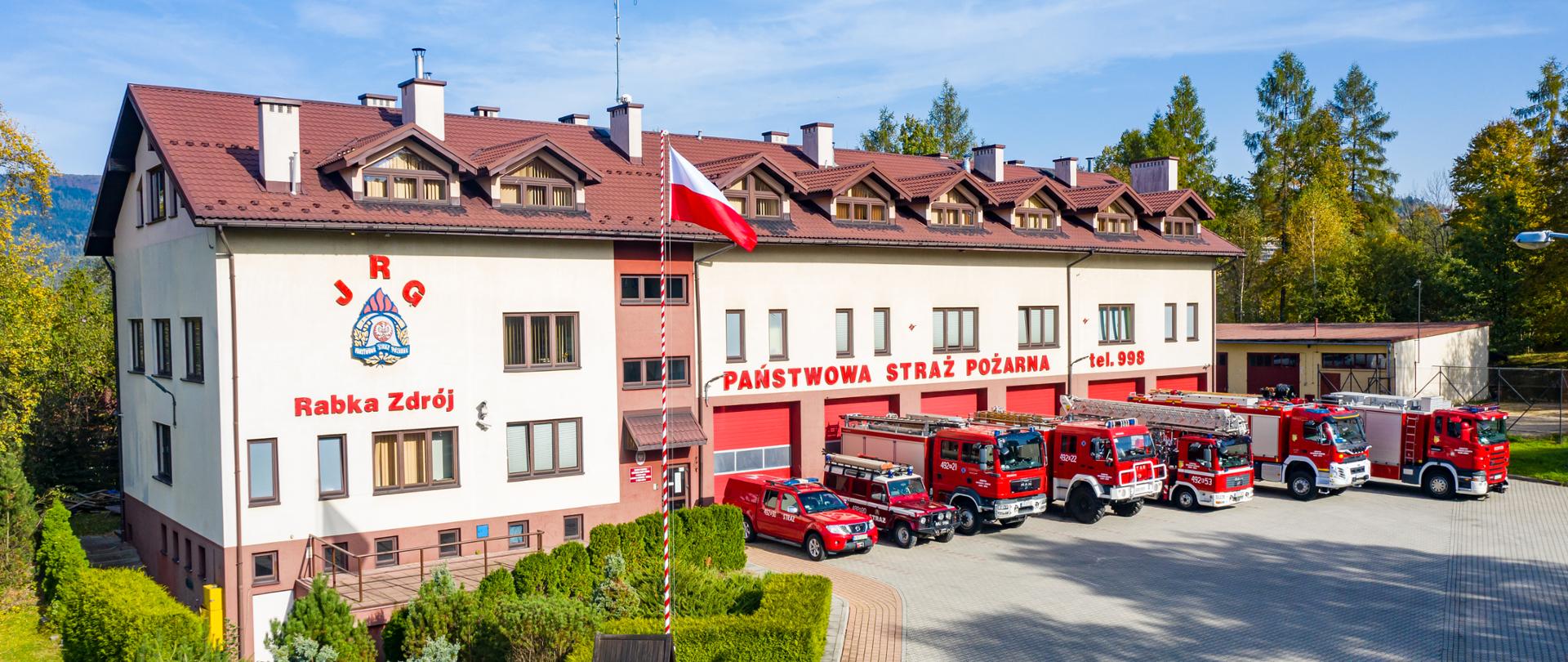 Zdjęcie budynku Jednostki ratowniczo-gaśniczej w Rabce-Zdroju. Na tle budynku powiewa flaga państwowa. Przed garażami zostały wystawione pojazdy pożarnicze, którymi dysponuje jednostka. Pojazdy są w kolorze czerwonym, i są to trzy samochody ratowniczo-gaśnicze, jeden samochód ratownictwa drogowego, jeden samochód podnośnik i dwa małe samochody terenowe. Wokół budynku znajdują się zagospodarowane tereny zielone: drzewa i krzewy. Na budynku znajdują się czerwone napisy: JRG Rabka-Zdrój z logo Państwowej Straży Pożarnej i napis Państwowa Straż Pożarna i tel. 998.