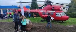 Na zdjęciu widoczny prawy bok czerwonego śmigłowca, przed którym przy głazie z tablicą stoi grupka dzieci. Jedno z dzieci podąża w stronę śmigłowca spoglądając na trzymaną w dłoni kartkę. W tle jasny budynek straży pożarnej, samochód strażacki. Na terenie zielonym przy czerwonym namiocie duża grupa osób. Niebo zachmurzone.