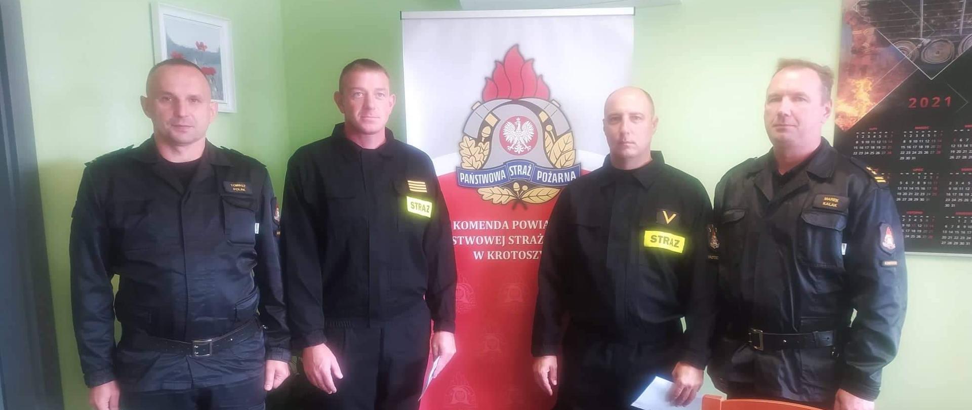 W gabinecie Komendanta Powiatowego PSP w Krotoszynie Komendant Powiatowy wraz ze swoim zastępcą gości dwóch strażaków, którzy wrócili z misji na terytorium Grecji. Zdjęcie podsumowujące spotkanie. Strażacy stoją przy banerze Komendy Powiatowej, tuż obok kierownictwo komendy. 