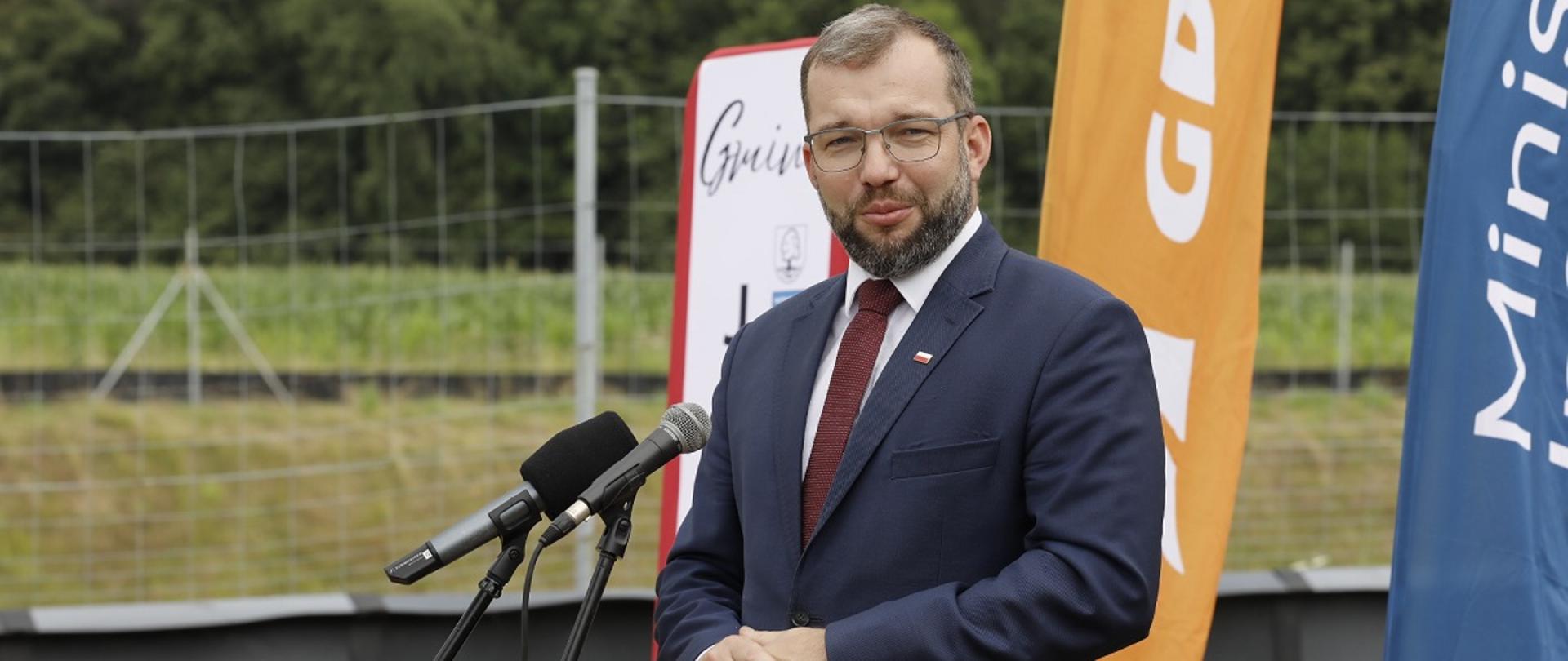 Minister funduszy i polityki regionalnej Grzegorz Puda stoi przy mikrofonie i przemawia, za jego plecami windery GDDKiA i Min. Infrastruktury