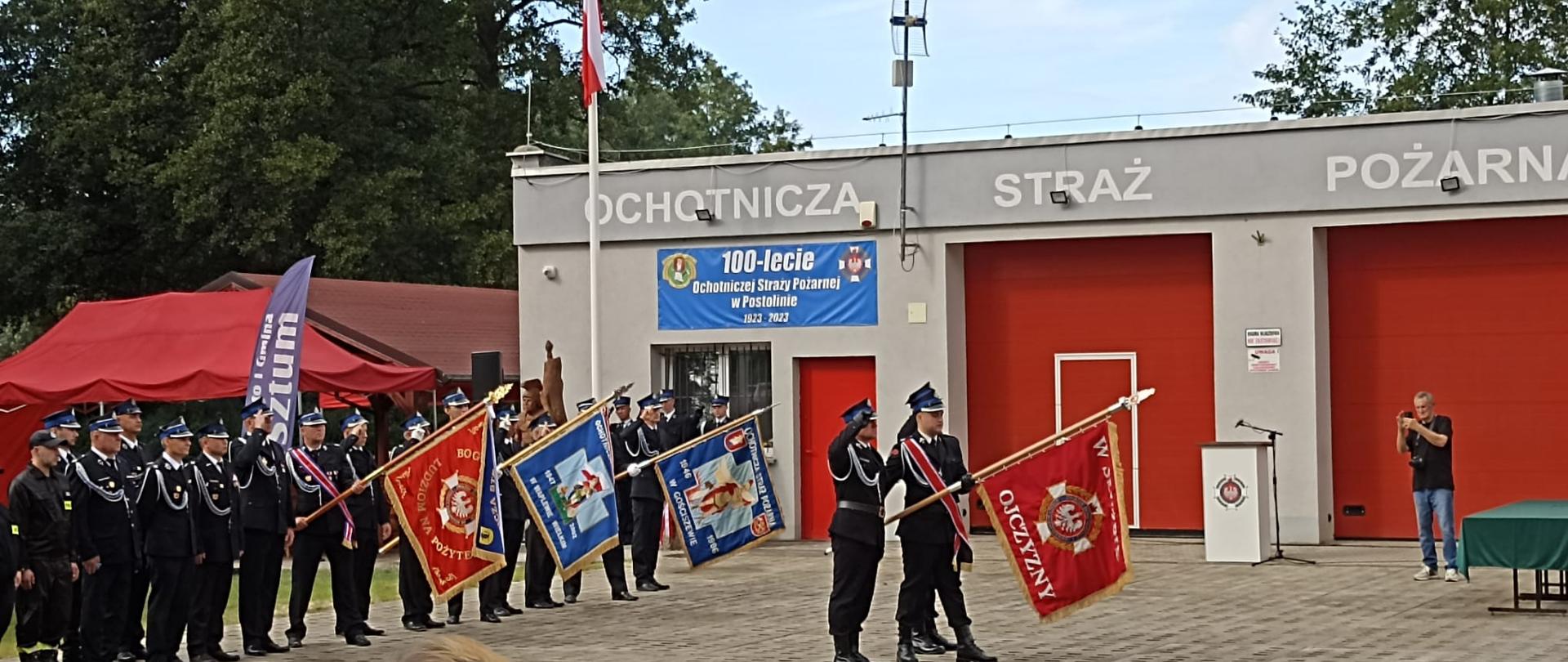 Przed remizą osp Postolin od lewej stoją: pododdziały reprezentacyjne Osp z terenu powiatu sztumskiego, przed nimi na środku zdjęcia poczet sztandarowy prezentujący nowo nadany sztandar.