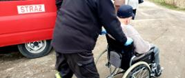 Starszy mężczyzna transportowany na wózku inwalidzkim przez strażaka. Przed nimi idą dwie osoby, z lewej strony fragment samochodu strażackiego.
