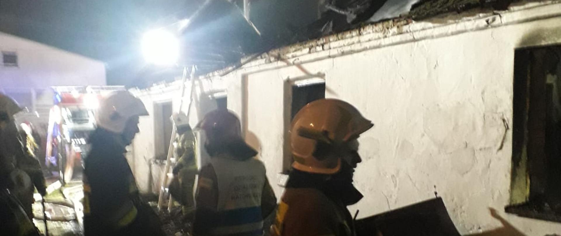 Zdjęcie przedstawia strażaków w trakcie działań. W tle znajduje się spalony budynek mieszkalny.