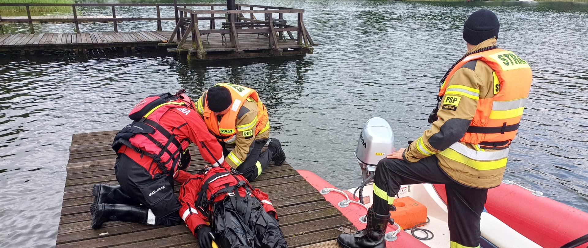 Na moście znajduje się trzech strażaków. Jeden z nich leży. Strażak, który leży ma na sobie czerwono-czarny kombinezon. Z lewej strony znajduje się strażak, który pochyla się nad leżącym strażakiem. Strażak ma na sobie czerwono-czarny kombinezon. Z prawej strony znajduje się strażak, który pochyla się nad leżącym strażakiem. Strażak ma na sobie umundurowanie w kolorze żółto-czarnym i pomarańczową kamizelkę ratunkową. Z prawej strony obok mostu znajduje się na wodzie łódź strażacka. Łódź jest koloru czerwono-białego. Na łodzi stoi strażak w umundurowaniu żółto-czarnym i pomarańczowej kamizelce ratunkowej. Strażacy maja założone czarne czapki. Wokół znajduje się jezioro. W tyle widać kolejny mostek oraz krzewy.