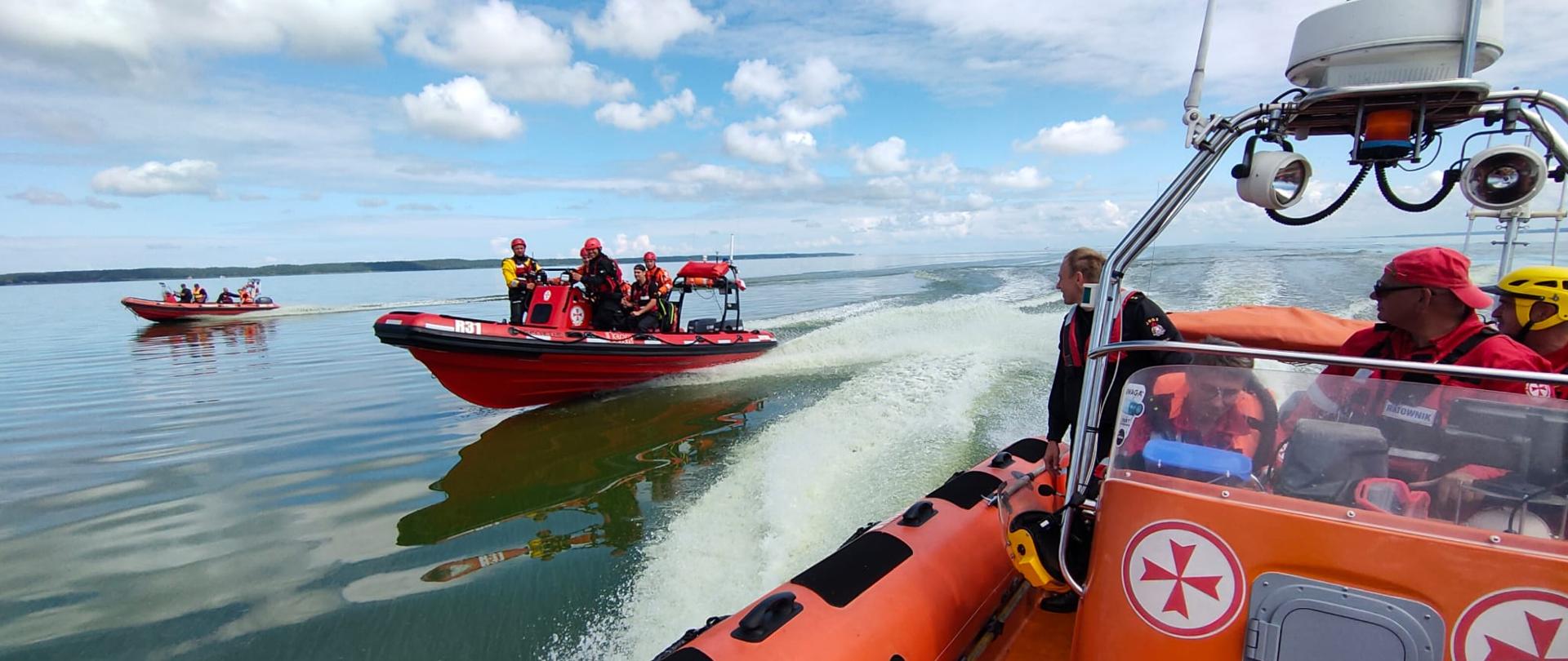 Trzy pomarańczowe łodzie motorowe płyną przez wody Zalewu Wiślanego. Na statkach znajdują się nurkowie państwowej straży pożarnej oraz służby morskiej.