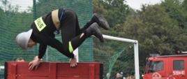 Na zdjęciu widać strażaka pokonującego w widowiskowym stylu ścianę pożarniczą. W tle widoczni obserwatorzy ćwiczeń oraz samochód pożarniczy. 