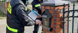 Strażak OSP trzymając ulotkę w ręku podchodzi do skrzynki pocztowej i umieszcza ulotkę w skrzynce pocztowej