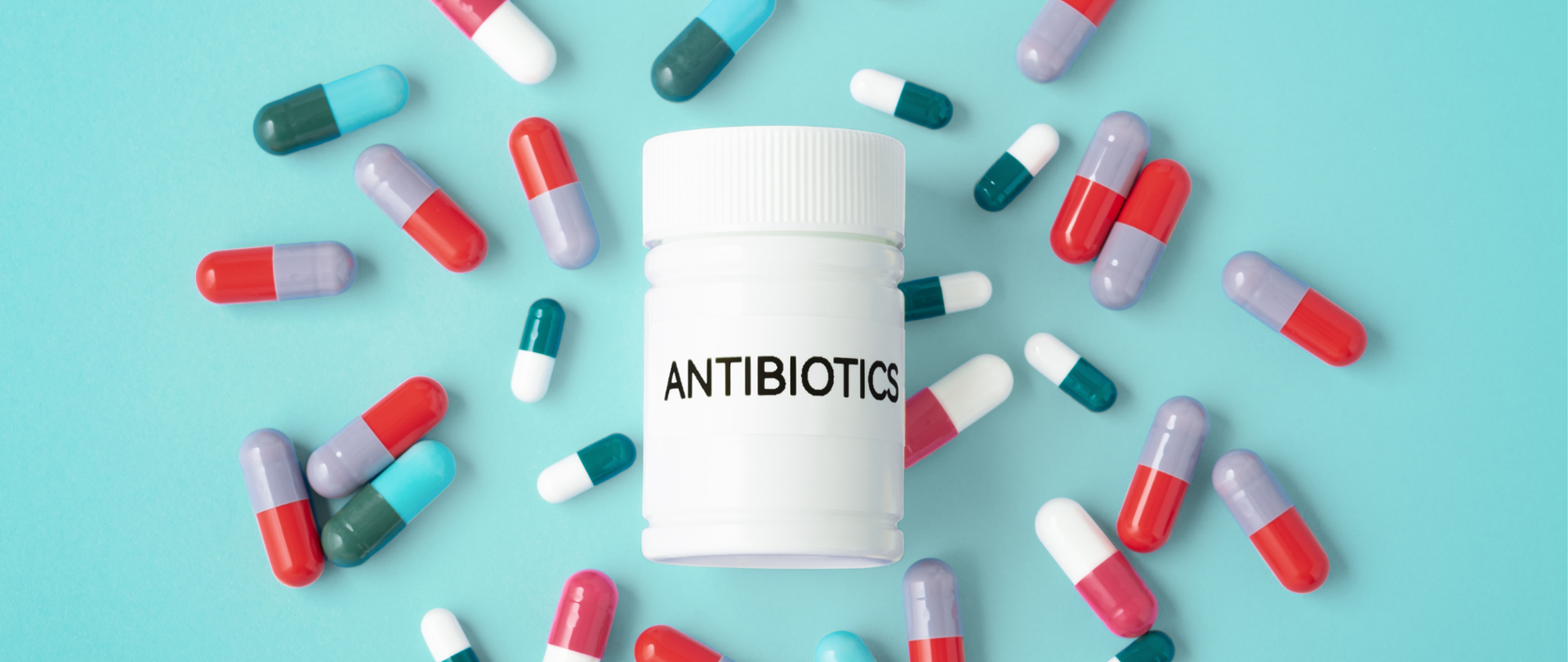 Zdjęcie przedstawia białą plastikową butelkę z napisem ‘ANTIBIOTICS’, ustawioną na środku błękitnego tła. Wokół niej rozsypane są kolorowe kapsułki i tabletki – czerwono-niebieskie, czerwono-fioletowe oraz biało-zielone. Całość tworzy kompozycję sugerującą temat stosowania antybiotyków.