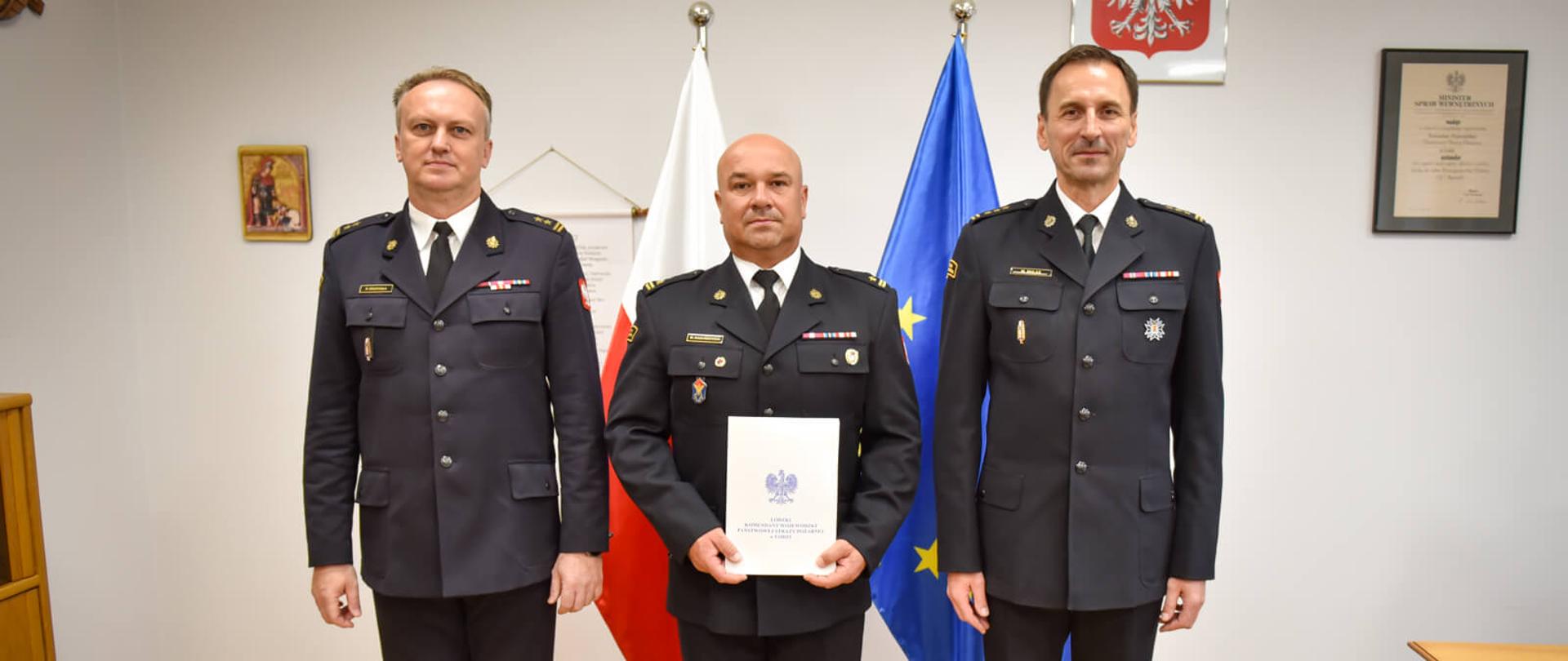 Oficer PSP w centrum trzyma w rękach dokument mianowania i pozuje do pamiątkowego zdjęcia wraz z dwoma przełożonymi. Za ich plecami widoczne są flagi Polski i Unii Europejskiej oraz godło państwowe.