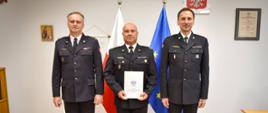Oficer PSP w centrum trzyma w rękach dokument mianowania i pozuje do pamiątkowego zdjęcia wraz z dwoma przełożonymi. Za ich plecami widoczne są flagi Polski i Unii Europejskiej oraz godło państwowe.