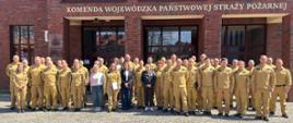 Grupa około 40 funkcjonariuszy w piaskowych mundurach oraz kilka osób w strojach cywilnych pozuje do wspólnego zdjęcia przed budynkiem z czerwonej cegły. Nad wejściem widnieje złoty napis: „Komenda Wojewódzka Państwowej Straży Pożarnej”. Wszyscy stoją na placu wyłożonym kostką brukową w pełnym słońcu.